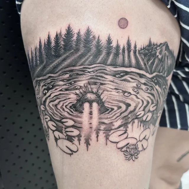 Tatovering av en innsjø med vannliljer, omgitt av en skog i bakgrunnen og en rund måne på himmelen, med et mystisk vesen som stikker hodet opp fra vannet.
