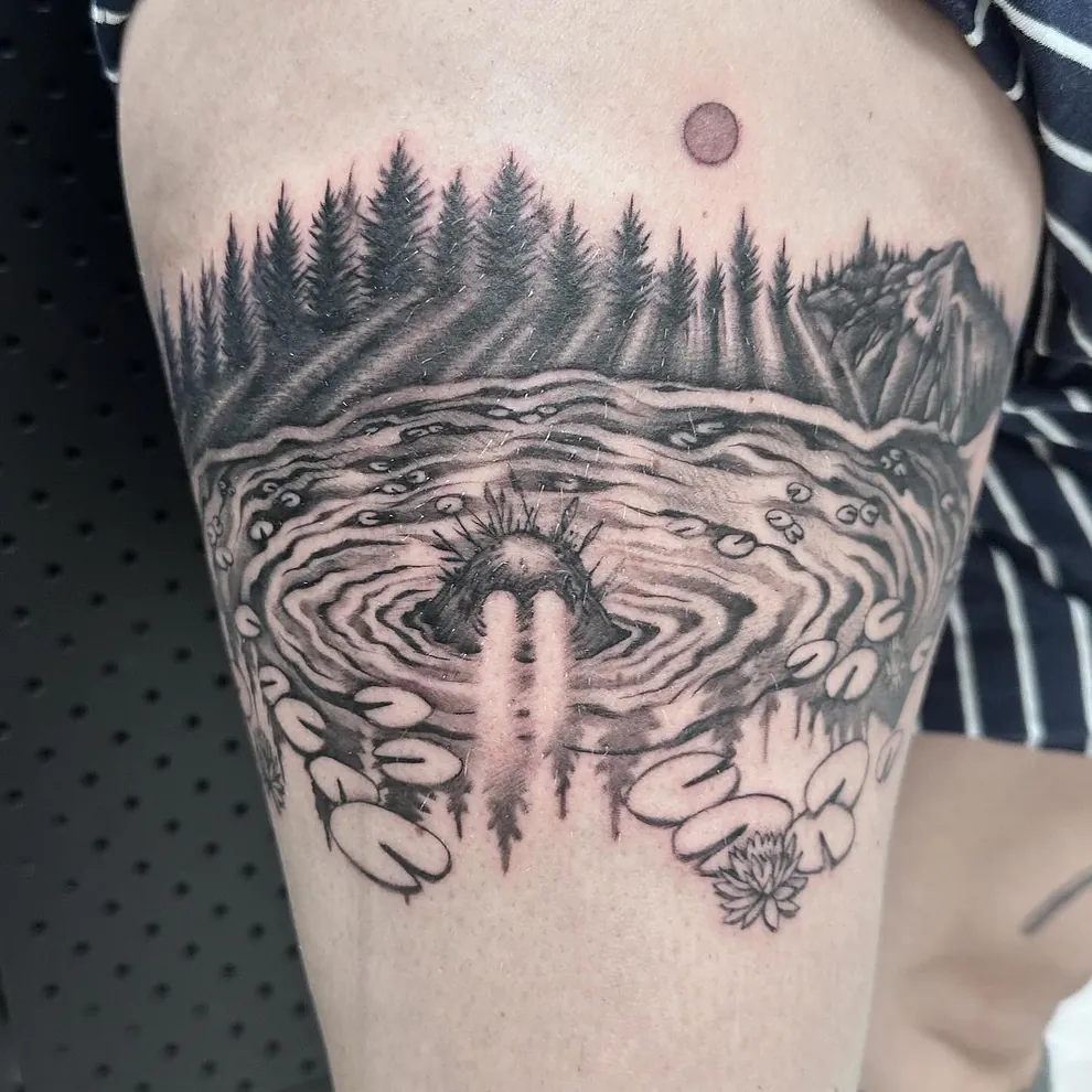 Tatovering av en innsjø med vannliljer, omgitt av en skog i bakgrunnen og en rund måne på himmelen, med et mystisk vesen som stikker hodet opp fra vannet.