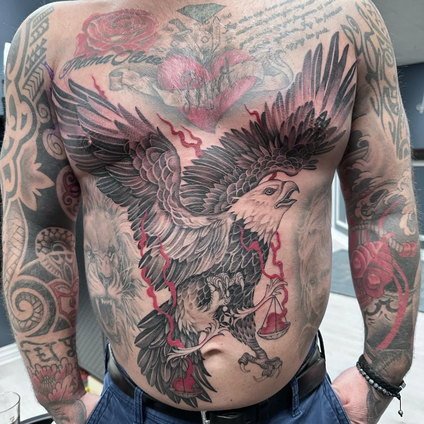 Stor brysttatovering av en ørn med spredte vinger og detaljer i rødt, som holder en vekt i klørne, omgitt av andre mindre tatoveringer.