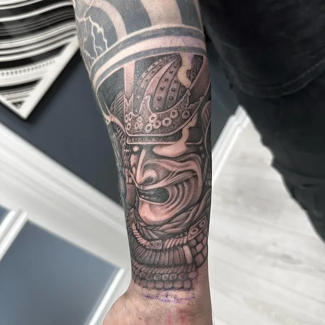 Tatovering på underarmen av en samurai-maske med dramatiske detaljer i svart-hvitt som gir et intens uttrykk.