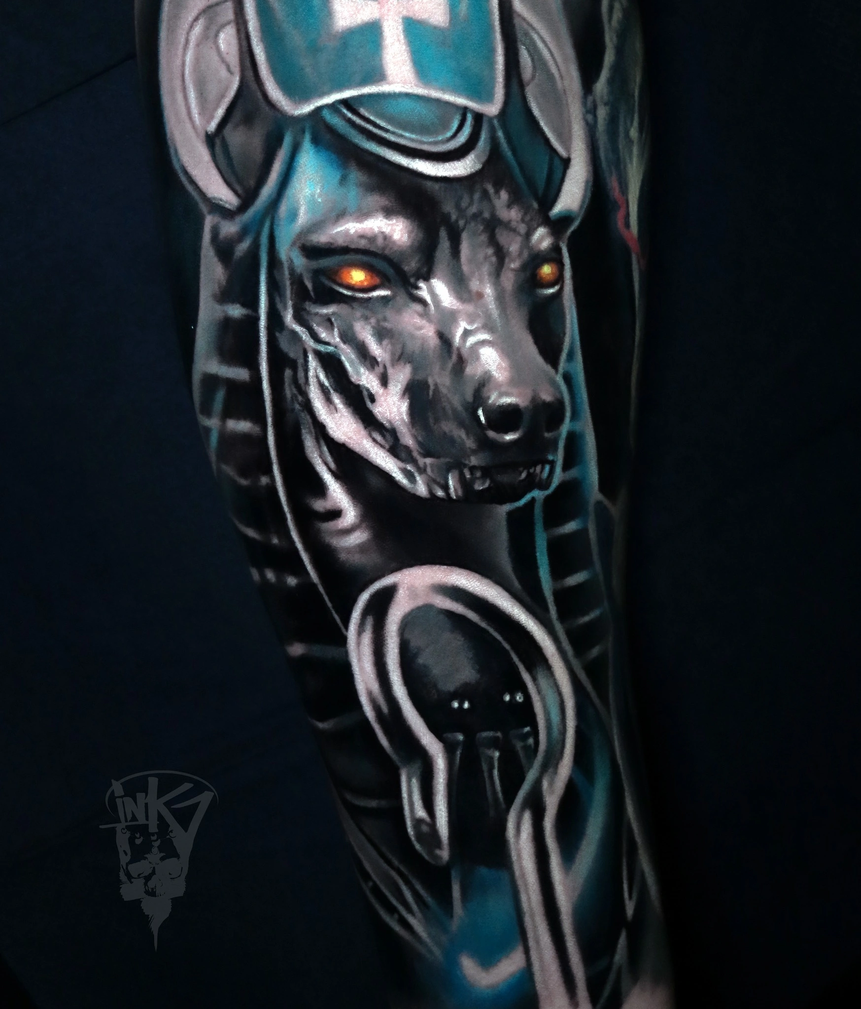 Tatovering av Anubis, den egyptiske guden med sjakals hode, gjengitt i mørke og lyse nyanser med glødende oransje øyne, som gir et kraftfullt og mystisk uttrykk.