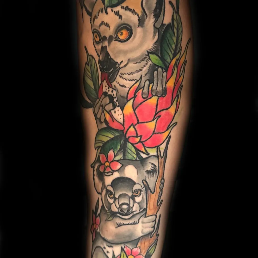 Fargerik tatovering av en lemur og en koala blant grønne blader og fargerike blomster, hvor lemuren holder et stykke frukt og ser fremover med store, gule øyne.