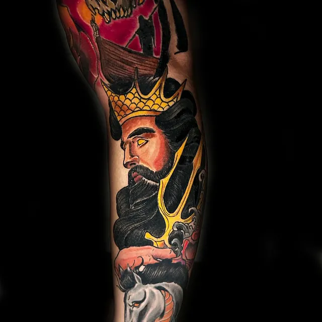 Tatovering på armen av en konge med mørkt hår og skjegg, iført en gullkrone, mens en hest og et skip med røde seil vises over hodet mot en dramatisk bakgrunn i rødt.