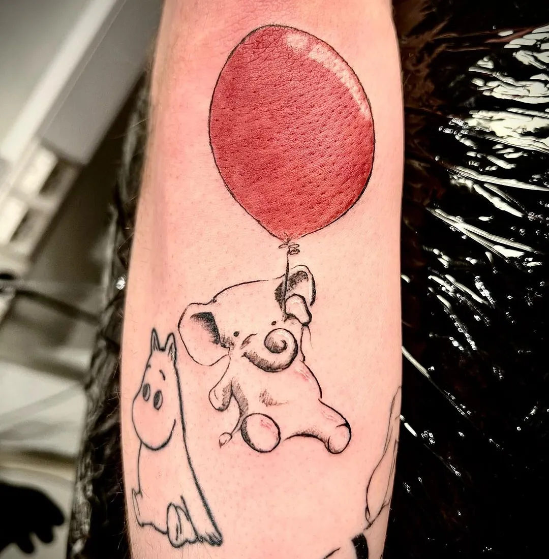 Enkel tatovering av en liten elefant som svever ved hjelp av en stor, rød ballong, med et skisset motiv av en Mummitrollet-karakter ved siden av.