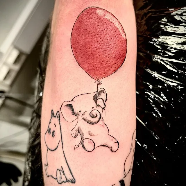 Enkel tatovering av en liten elefant som svever ved hjelp av en stor, rød ballong, med et skisset motiv av en Mummitrollet-karakter ved siden av.